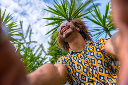 Afro-haired man on summer vacation next to some palm trees next to the beach taking a selfie with both hands smiling. Travel and tourism conceptの写真素材