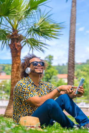 Afro haired man sitting on the grass on summer vacation next to some palm trees by the beach. Traveling and tourism concept, listening to music with headphonesの写真素材