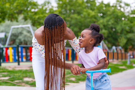 Black African ethnicity mother having fun with her daughter in playground walking with the skateboard and sticking out her tongueの写真素材