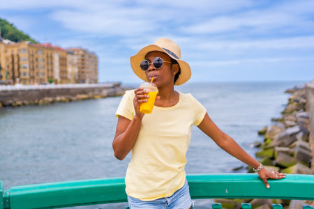 Black ethnic woman having an orange juice by the sea, tourist enjoying the summerの写真素材