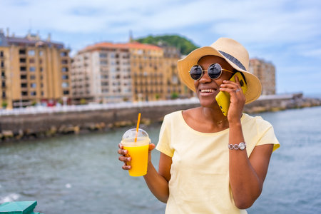 Black ethnic woman drinking orange juice texting with phone, tourist enjoying summerの写真素材