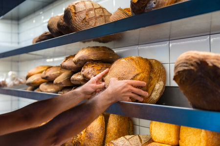Hands of baker employee of bakery in workshop workshop of artisan bakery placing baked bread from oven on shelfの写真素材