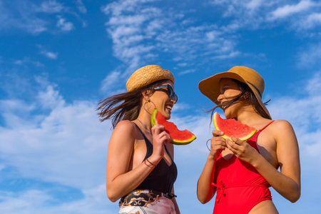 Female friends at the beach in summer eating a watermelon with the sky in the background, shot from...