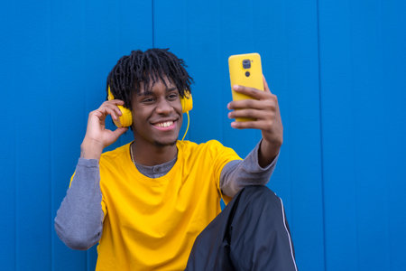 Happy afro man taking a selfie while listening to music sitting on the floor next to a blue backgroundの写真素材