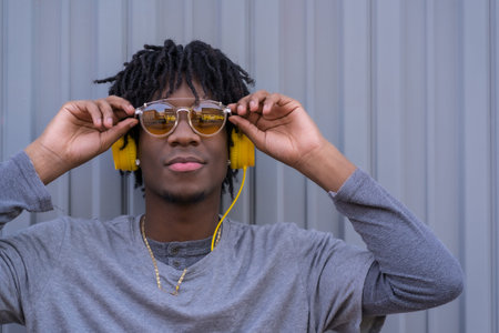 Confident african young man with sunglasses and headphones standing next to an urban gray background
