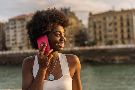 Close-up portrait of a distracted and smiling african young woman using phone next to the sea during sunsetの写真素材