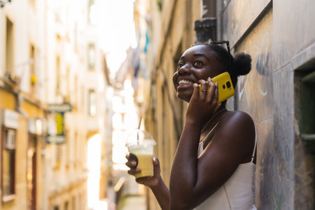 Afro woman leaning on a city wall drinking fresh juice and talking to the mobileの写真素材