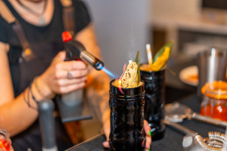 Cropped photo with close-up of a female bartender flaming the garnish of a cocktail with a torch on the counter of a barの写真素材