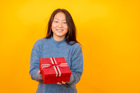 Studio photo with yellow background of a chinese woman giving a present smiling at cameraの写真素材