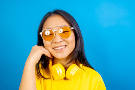 Studio photo with blue background of a shy and cute chinese woman wearing sunglasses smiling at cameraの写真素材