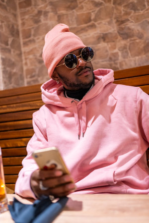 Vertical portrait of a cool african man wearing pink clothes using phone sitting in a barの写真素材