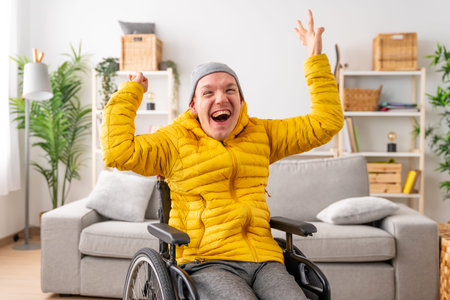 Portrait of a happy disabled man in wheelchair raising hands in joy celebrating in the living room...