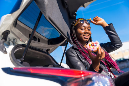 African woman enjoying a donut sitting in the trunk of a car on a sunny dayの写真素材