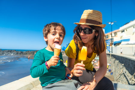 A woman and a child are sitting on a wall near the ocean, eating ice cream. The woman is wearing a straw hat and sunglassesの写真素材