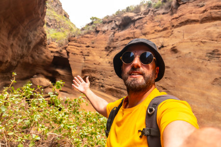 A man in a yellow shirt and sunglasses is taking a selfie in front of a rocky cliff. Concept of adventure and exploration, as the man is posing in a natural setting with a backpack onの写真素材