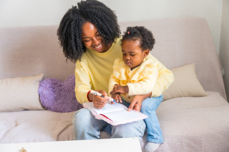 Little african daughter drawing sitting on the lap of her mother on the sofa in the living room at homeの写真素材