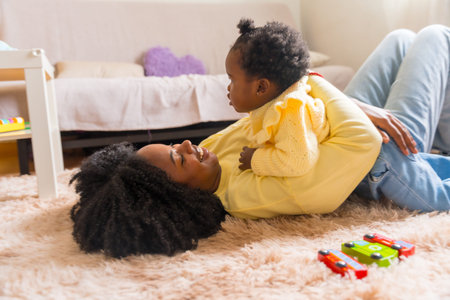 Playful and smiling african woman lying on the floor embracing her daughter in the living roomの写真素材