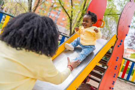 Happy african child playing with her mother on the slide in a public playground