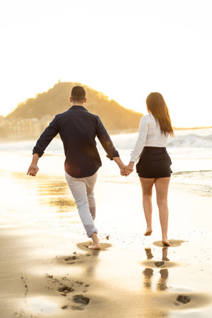 Vertical photo of the rear view of a playful couple running holding hands on the edge of the sea during sunsetの写真素材