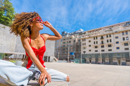 Horizontal photo with copy space of a beauty latin young fashionable woman sitting in an urban square enjoying sun wearing heart shape sunglassesの写真素材