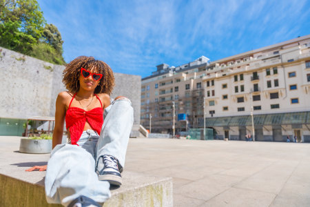Latin young woman with heart shape sunglasses sitting on the ground outdoors enjoying summer in the cityの写真素材