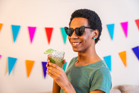 Portrait of a happy latin man with sunglasses drinking cocktail next to garlands decorating a house during celebrationの写真素材