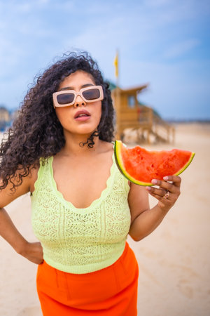 A woman is holding a watermelon on a beach. She is wearing a green tank top and orange skirt. The image has a bright and cheerful moodの写真素材