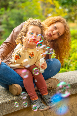 Vertical photo of a caucasian woman and daughter playing blowing bubbles in a park