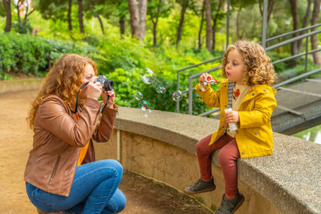 Caucasian adult woman taking a photo with a digital camera of her daughter blowing soap bubbles in a...