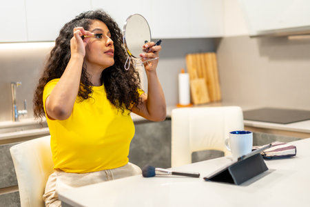 Latin beauty young woman applying makeup to eyelashes sitting in the kitchen at homeの写真素材
