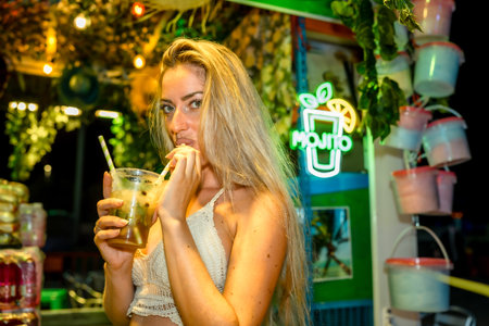 Sensual blonde woman drinking cocktail with straw at night in a bar with tropical decorationの写真素材