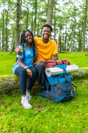 Vertical portrait of young african friends embracing sitting on a log together in a green forest in springの写真素材
