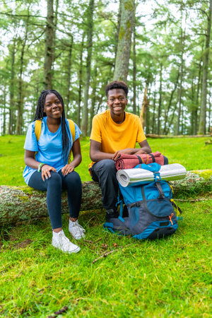 Vertical portrait of two young male and female african hikers resting on a log smiling surrounded by green treesの写真素材