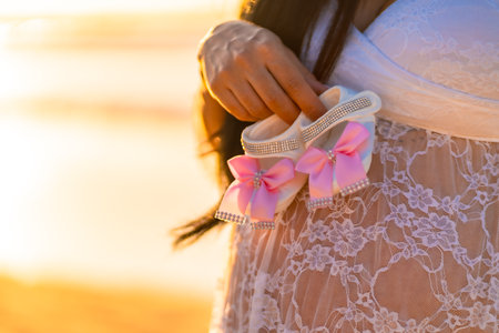 A woman holding a pair of pink and white baby shoes. The shoes are decorated with a bow and are being held in her hand. Concept of anticipation and excitement for the upcoming arrival of a new babyの写真素材