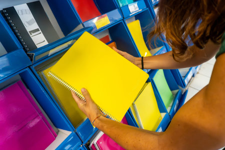 A woman is holding a yellow spiral notebook in a store. The notebook is on a shelf among other...
