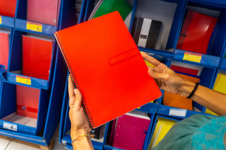 A person is holding a red spiral notebook in front of a shelf full of notebooks. The person is looking at the notebook, possibly considering which one to buyの写真素材
