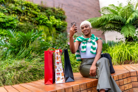 African woman taking selfie sitting next to colorful shopping bags outside a mall after shoppingの写真素材