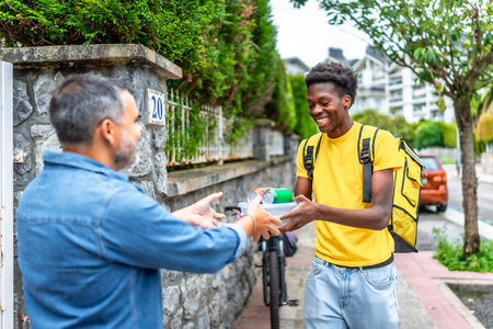 Mature man receives food at home from an African delivery person by bikeの写真素材