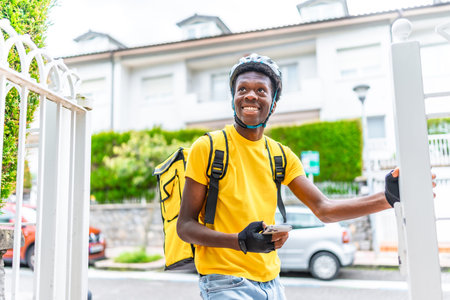 Cheerful african young delivery person on a bike delivering a package to a house, waiting on the...