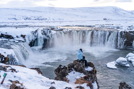 Woman hiker sitting looking at frozen and snowy Godafoss waterfall in winter in Icelandの写真素材