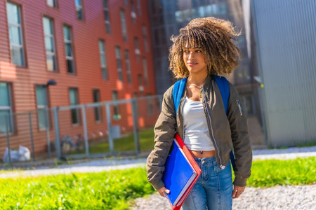 Confident latin young woman standing in the university campus looking away holding notebooks, folders and rucksackの写真素材