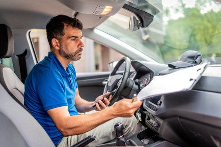 Delivery man using mobile phone and doing paperwork before delivering packages sitting inside a carの写真素材