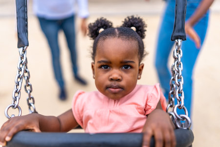 Cute african baby girl sitting on swing in a public park next to her familyの写真素材