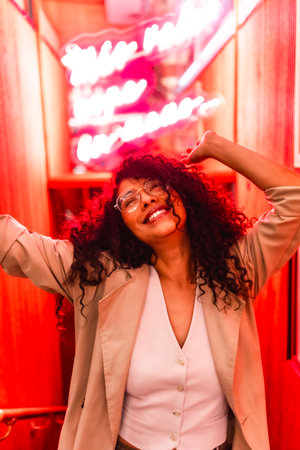 Young and happy latin beauty woman with curly hair dancing near red neon signの写真素材