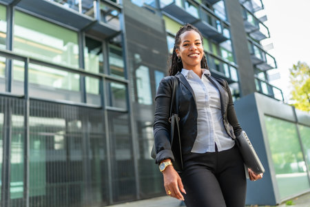 Confident latin modern businesswoman next to block office smiling at camera and carrying laptop and handbagの写真素材