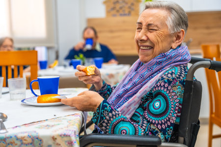 Side view of a happy elder woman with disability eating breakfast sitting on wheelchair at geriatricsの写真素材