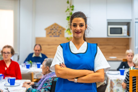 Portrait of a young confident female caregiver in blue apron standing proud at geriatrics dining room with senior people around having breakfastの写真素材