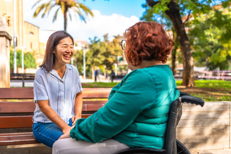 Happy and cheerful asian caregiver sitting on park bench talking with disabled woman sitting on electric wheelchair during sunny dayの写真素材