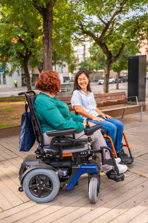 Vertical side view photo of two multi-ethnic women with disabilities in wheelchair talking relaxed in a parkの写真素材