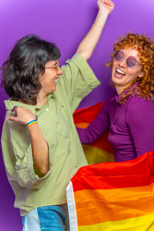 Studio portrait with purple background of two happy lesbian couple dancing wrapped with lgbt rainbow...